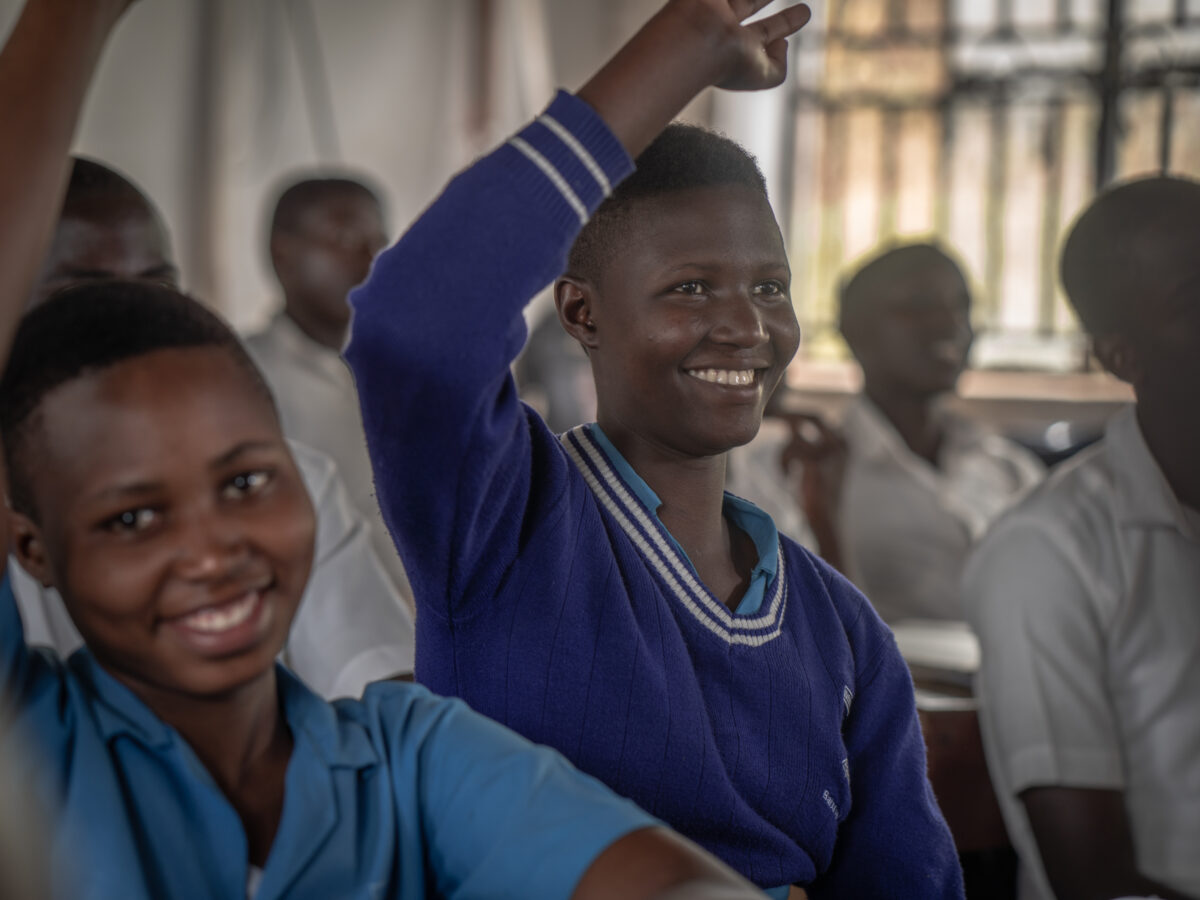A student in a Ugandan classroom raising her hand and smiling during a lesson