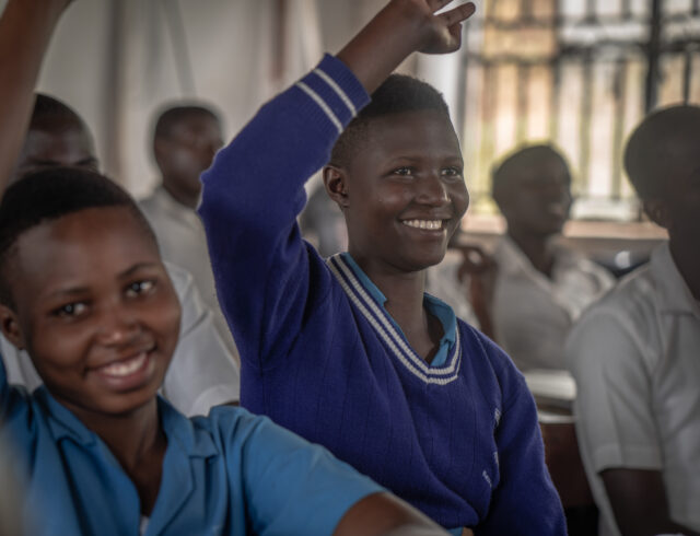 A student in a Ugandan classroom raising her hand and smiling during a lesson